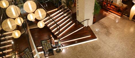 A modern hotel lobby with a wooden staircase, hanging spherical lights, and a reception desk.