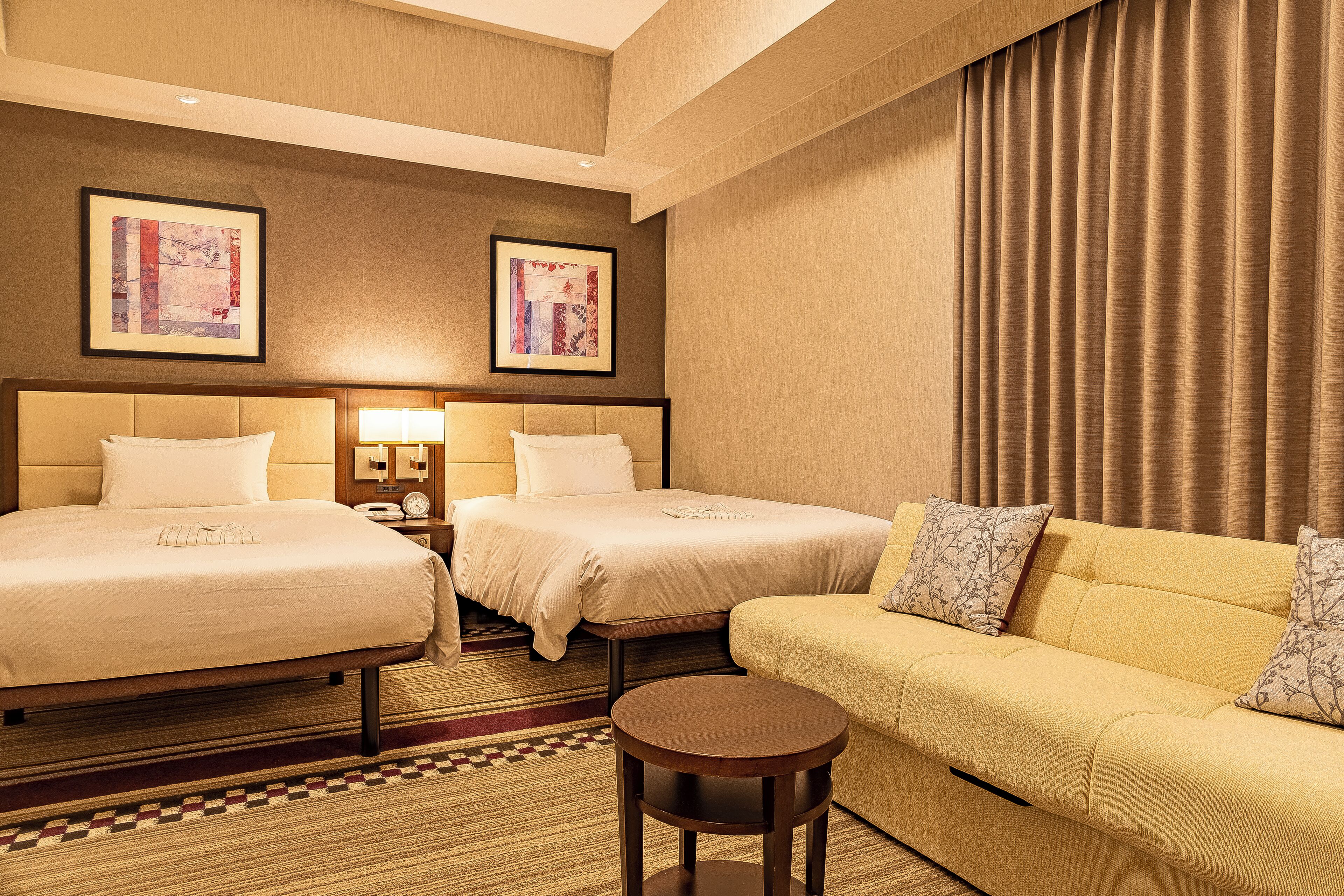 A hotel room with two beds, a sofa, a small table, and two framed artworks on the wall.
