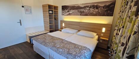 Habitación de hotel con una cama grande, cabecero de madera, mesitas de noche, una mesita auxiliar y una pintura de un paisaje urbano.