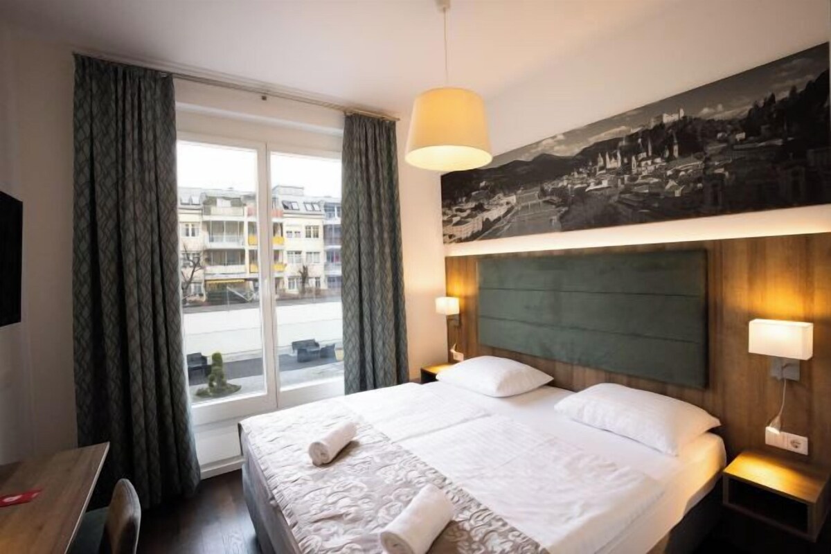 A hotel room with a large bed, two bedside lamps, a view of buildings through the window, and a painting on the wall.