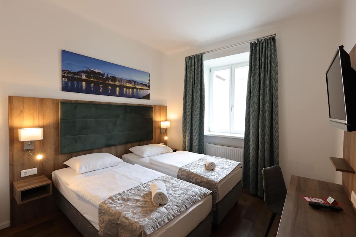 A hotel room with two beds, a wooden headboard, a painting of a cityscape, a TV, and a desk with a remote control.