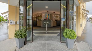 Entrada de hotel com porta de vidro, balcão de recepção e duas plantas em vasos.
