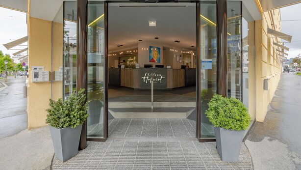 A hotel entrance with a glass door, a reception desk, and two potted plants.
