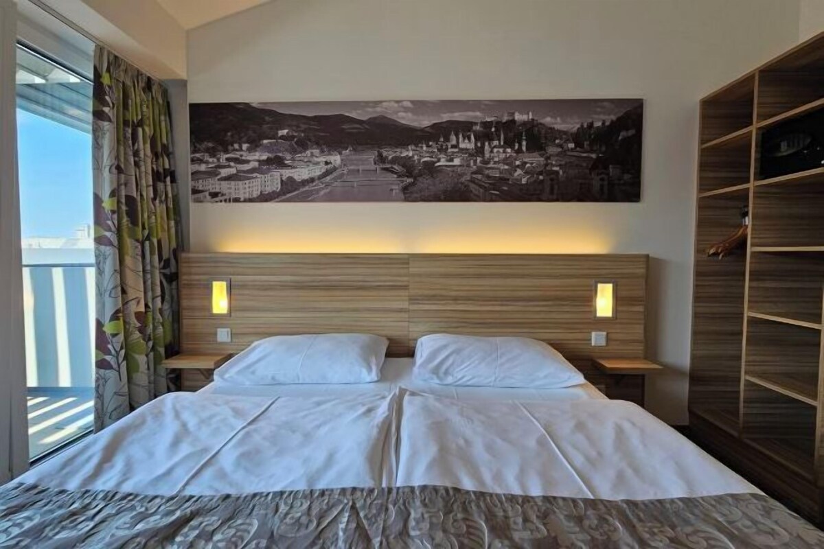 Ein Hotelzimmer mit einem großen Bett, einem Kopfteil mit zwei Wandleuchten, einem Stadtbild über dem Bett und einem Balkon mit Vorhängen.