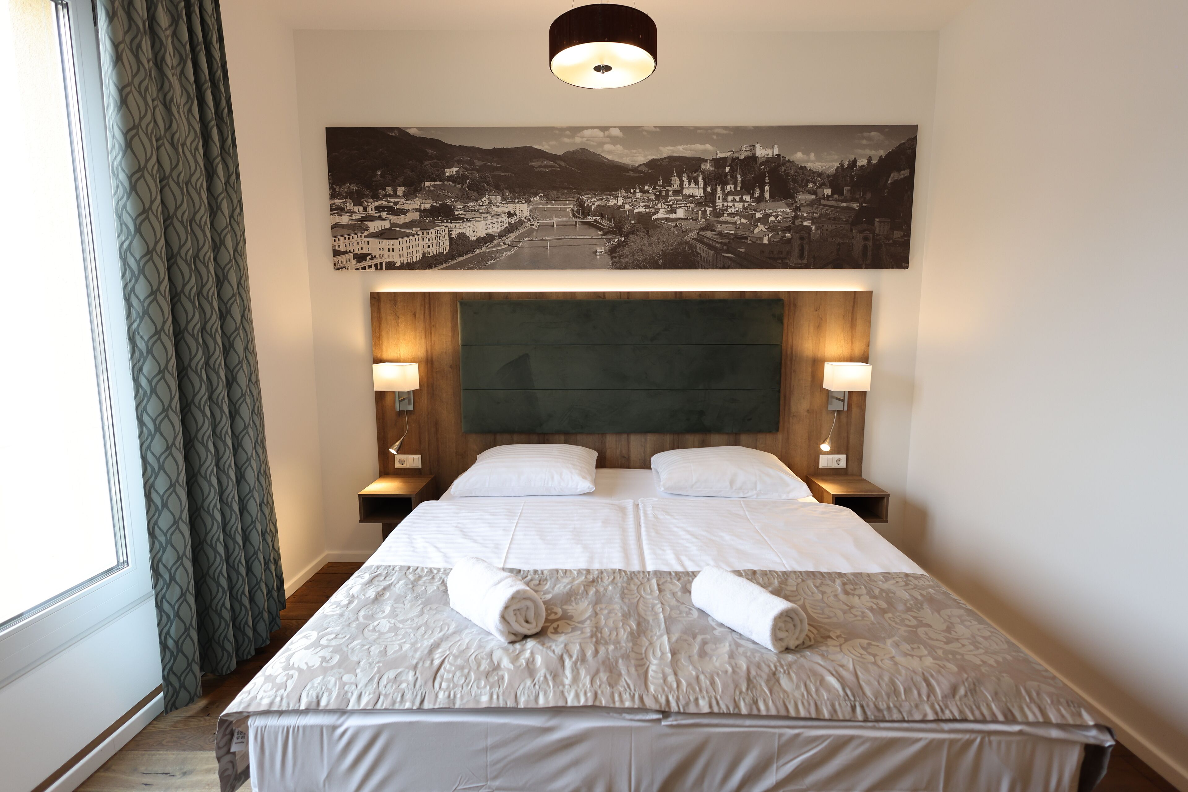 Una habitación de hotel con una cama grande, dos lámparas de noche, un cabecero y un cuadro de paisaje urbano sobre la cama.