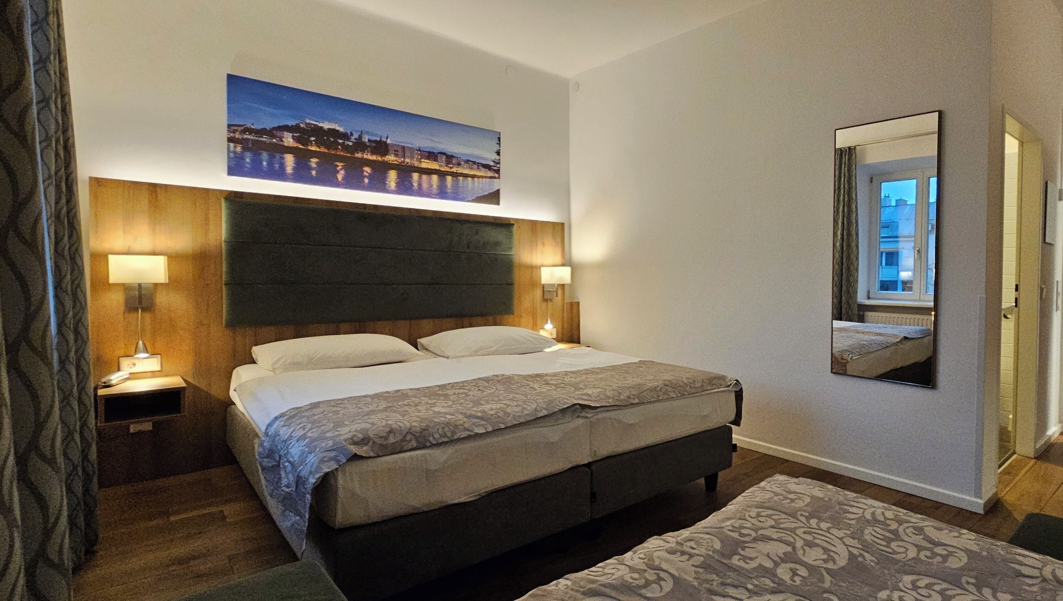 A hotel room with a bed, bedside lamps, a large mirror, and a cityscape painting on the wall.