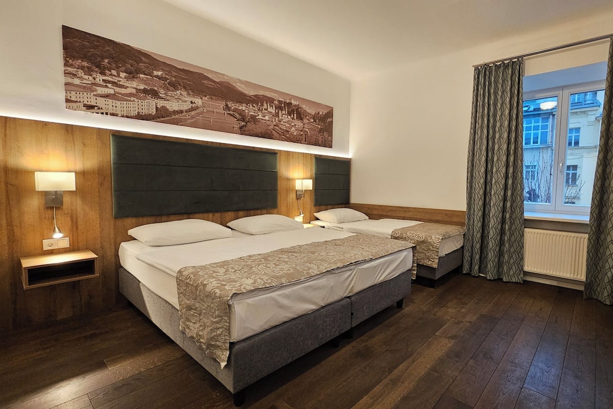 A hotel room with a large bed, two bedside tables, a wall-mounted lamp, a window with curtains, and a cityscape painting above the bed.