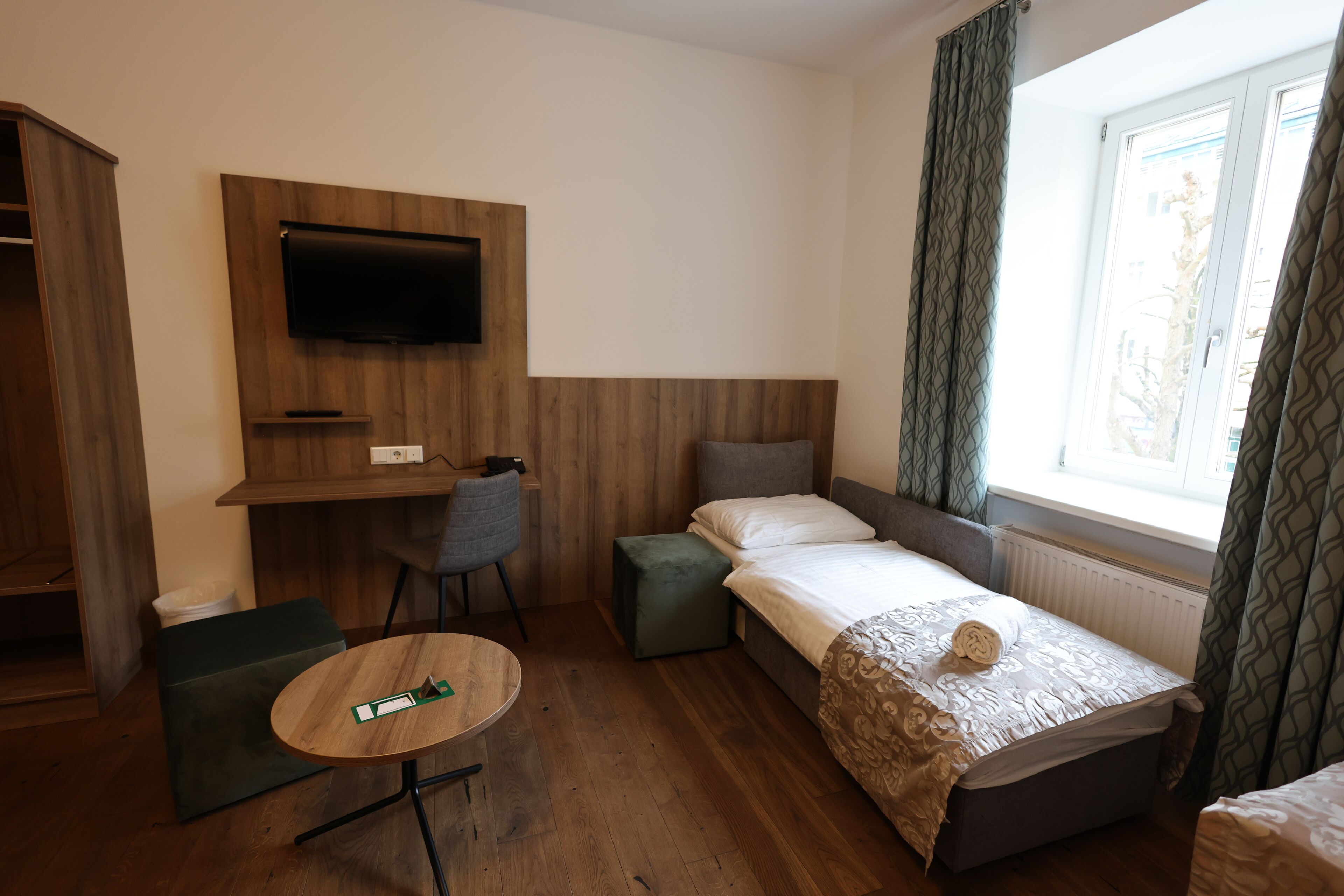 A compact hotel room with a bed, a desk, a chair, a small table, and a TV mounted on the wall.