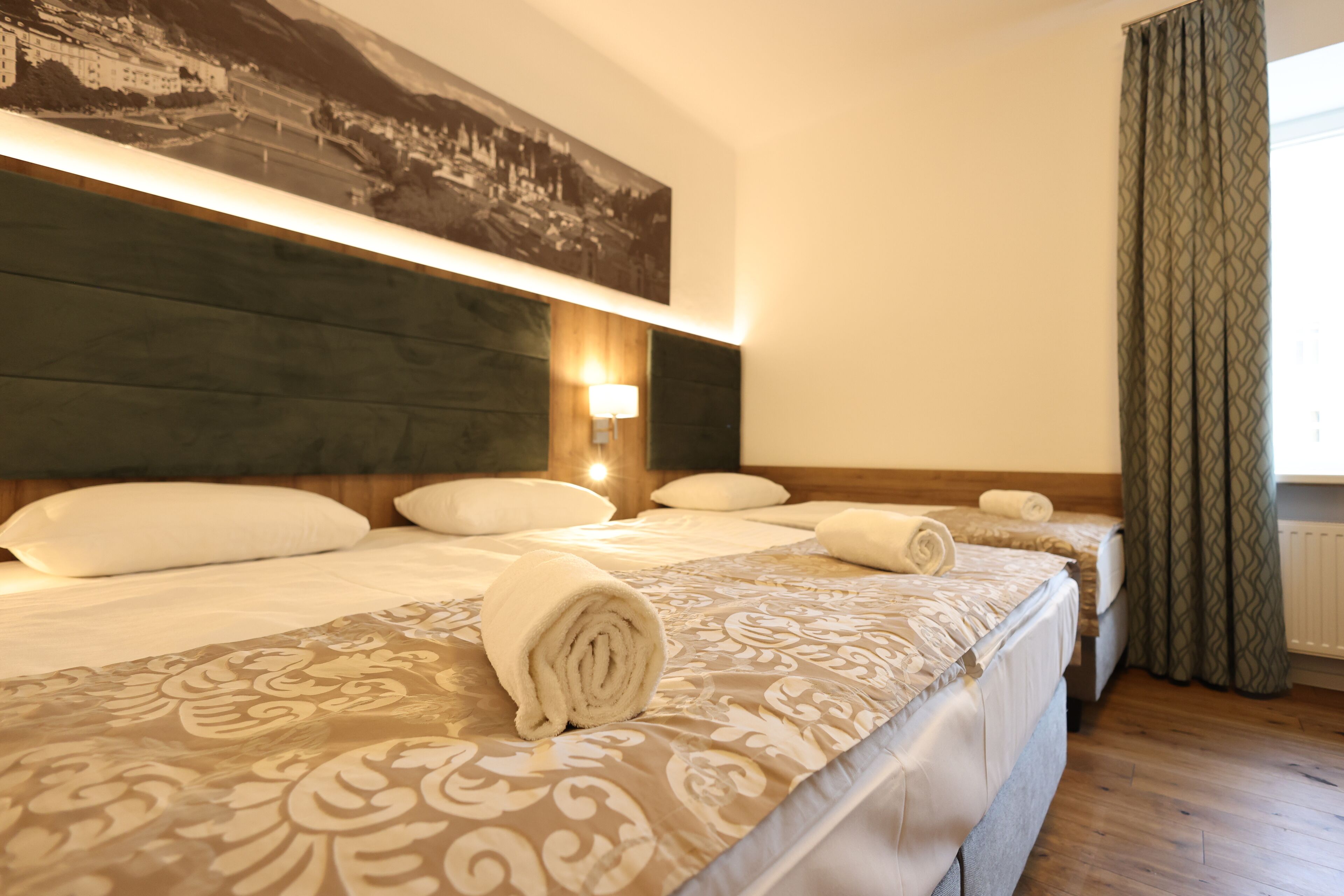 Una habitación de hotel con una cama grande, un cabecero con una pintura de paisaje urbano, una lámpara de pared y una ventana con cortinas.