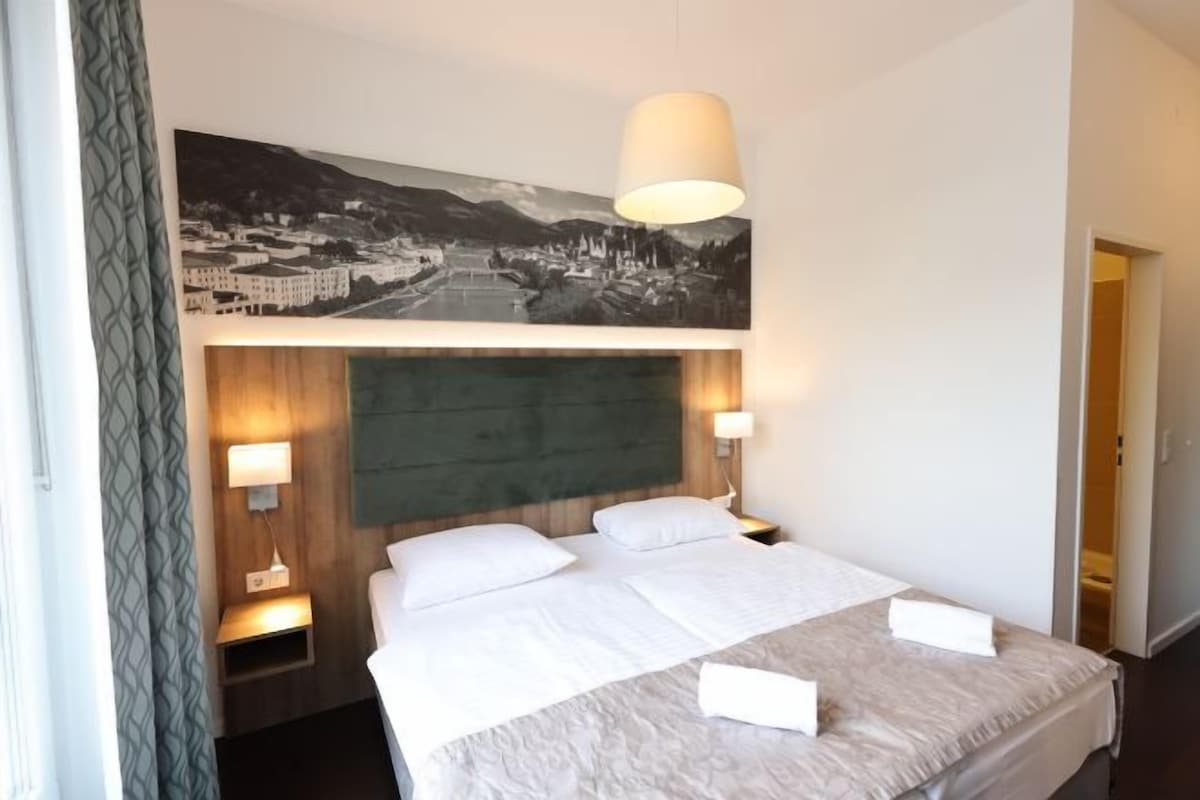 Hotellihuone, jossa on suuri sänky, kaksi yöpöytävalaisinta, sängynpääty ja seinälle ripustettu taulu.