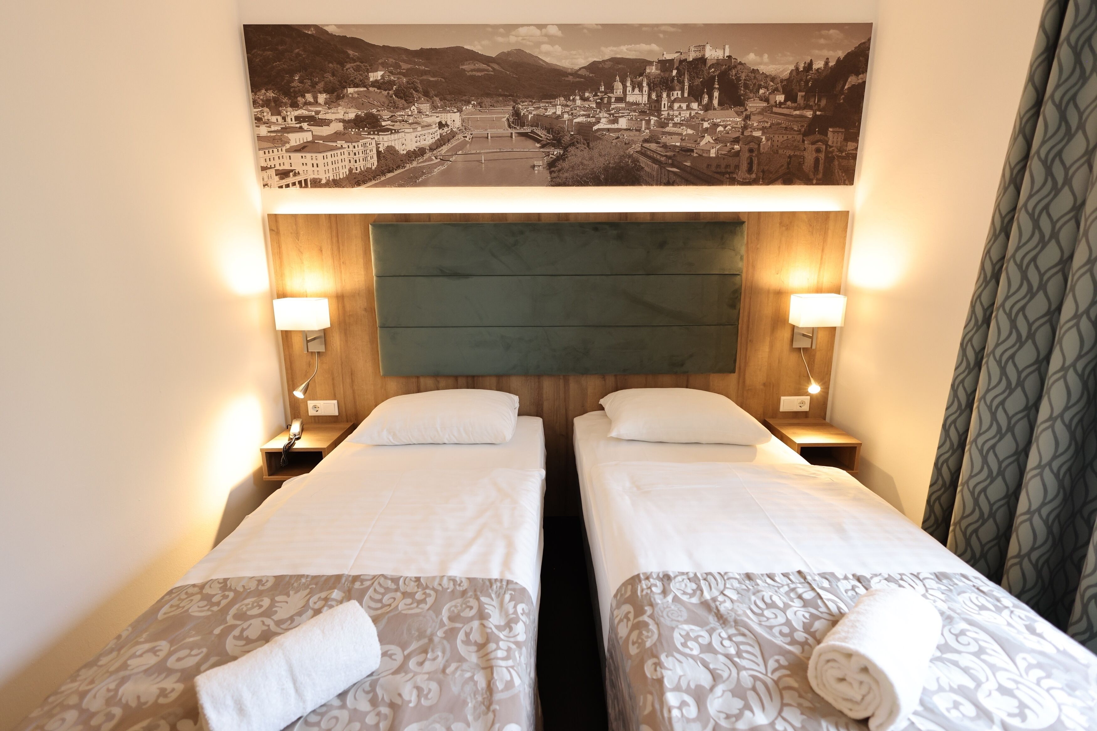 Une chambre d’hôtel avec deux lits simples, une grande photographie encadrée représentant un paysage urbain au-dessus des lits et des lampes de chevet.