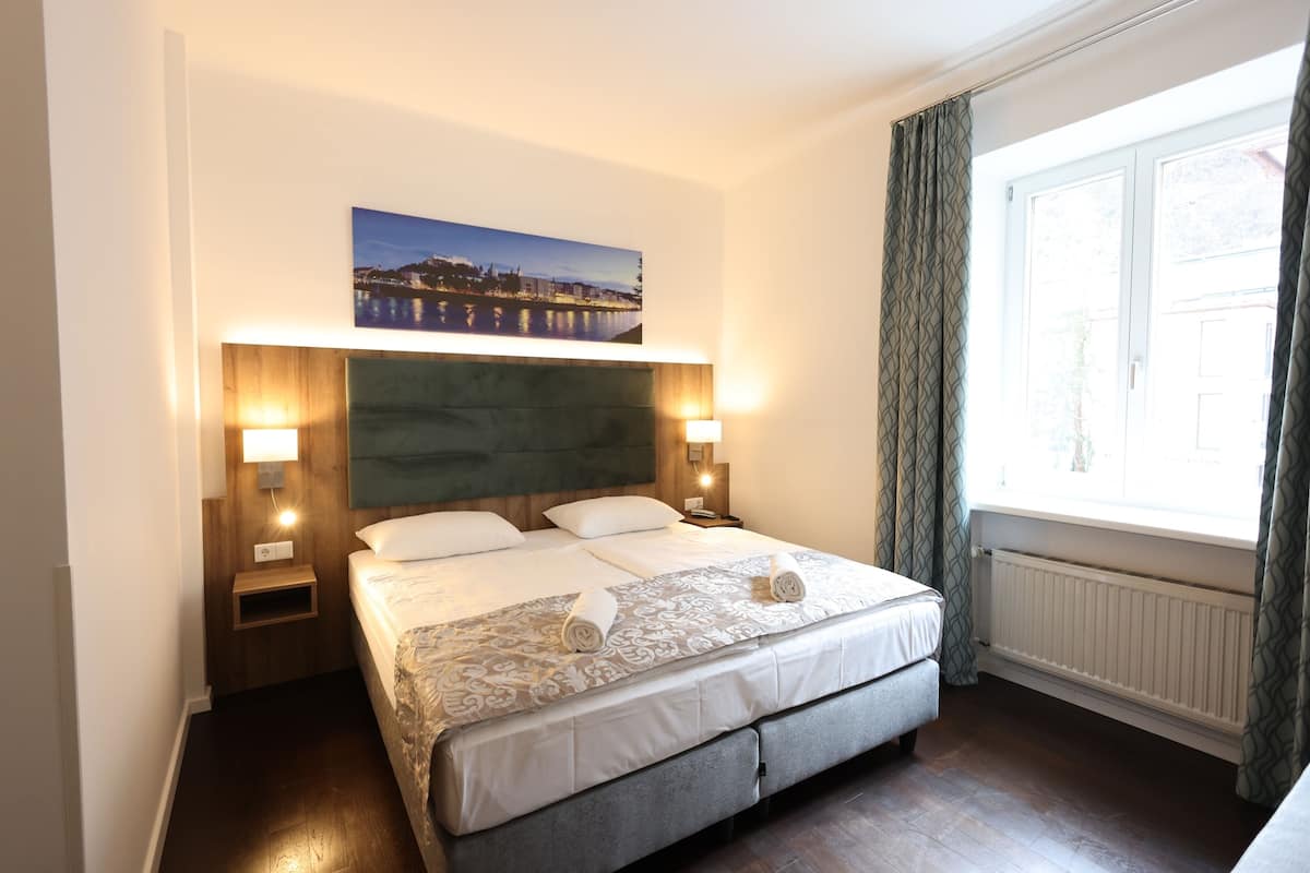 A hotel room with a large bed, two bedside tables with lamps, a headboard with a cityscape painting, a window with curtains, and a radiator.