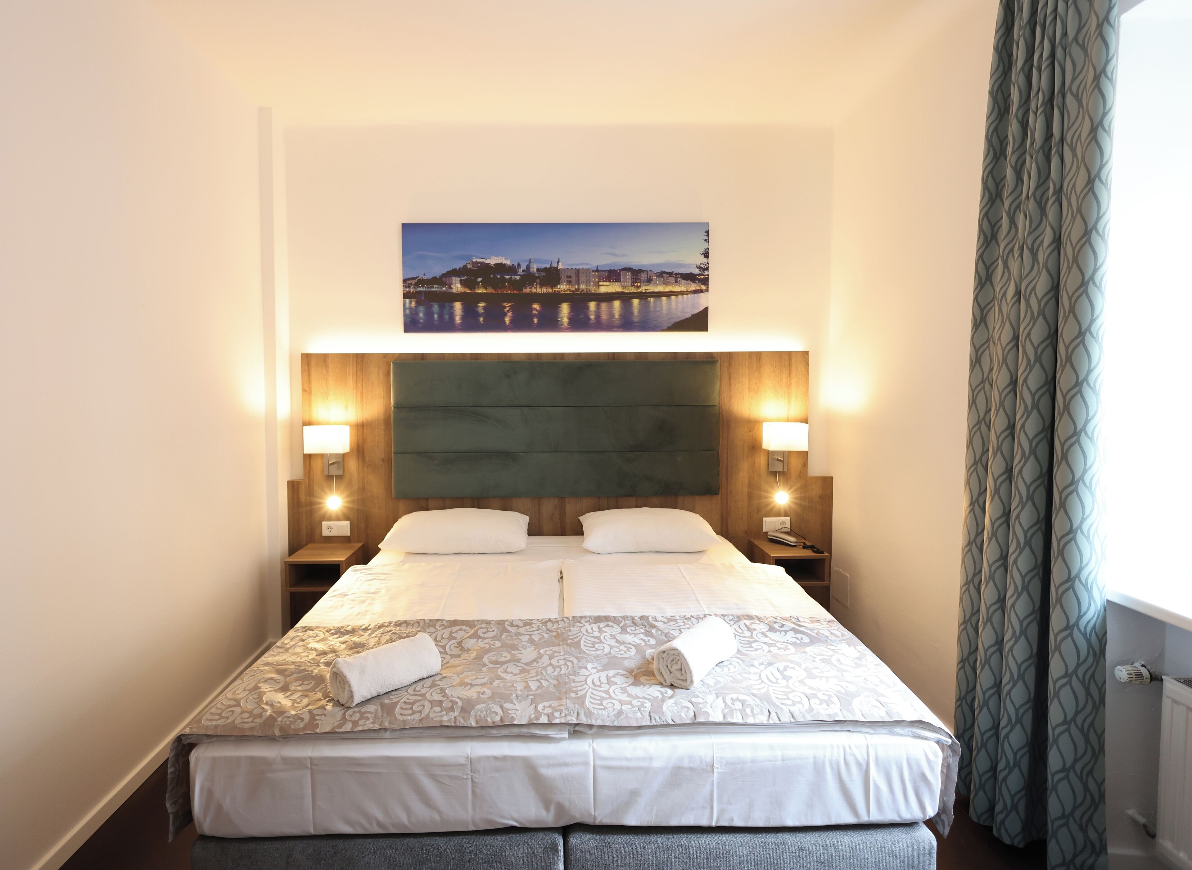 A neatly made bed with a patterned bedspread and two pillows, a headboard with a cityscape painting, two bedside lamps, and a view of a cityscape through a window with curtains.