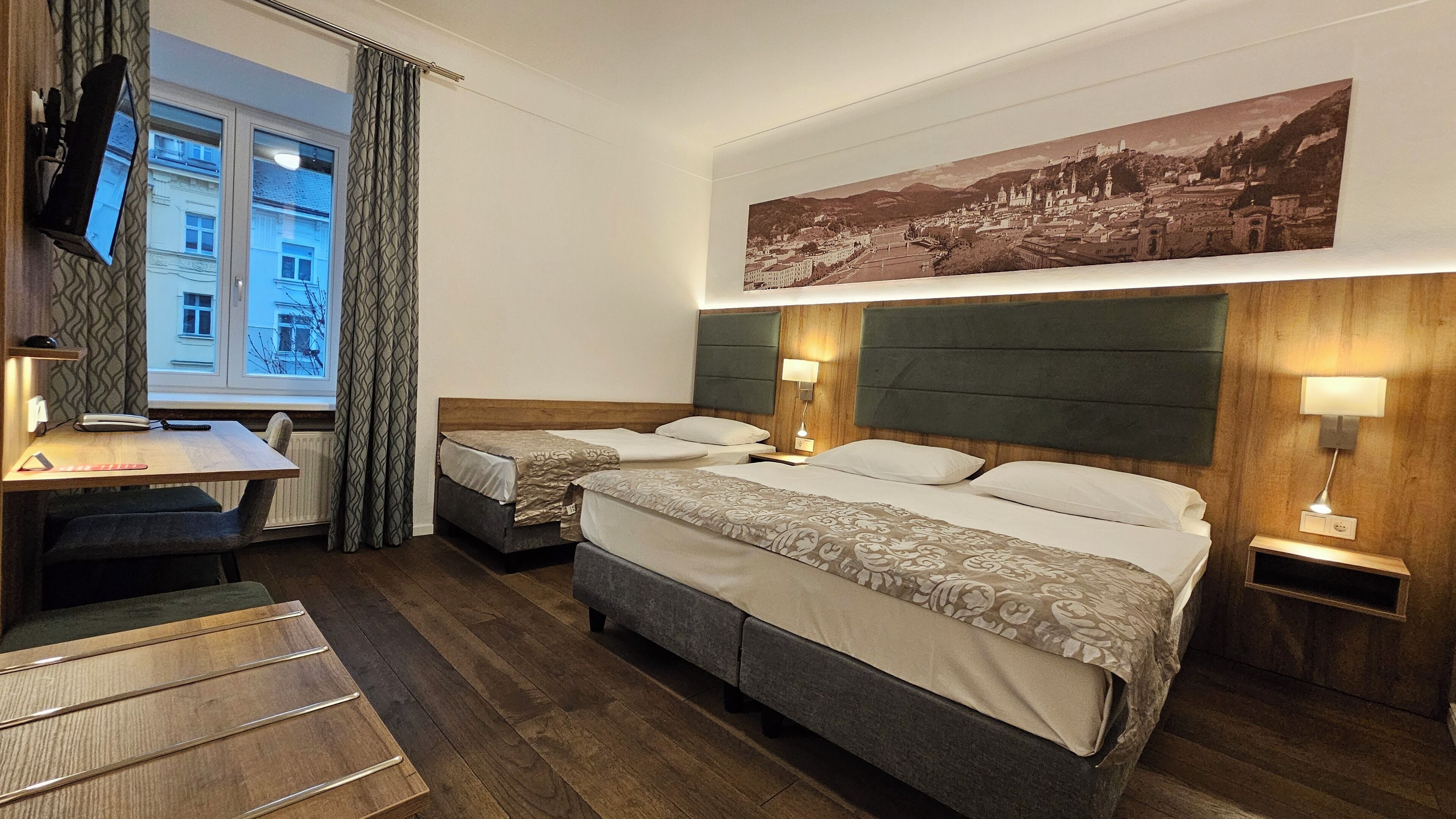A hotel room with two beds, a desk, a chair, a TV, and a cityscape painting.