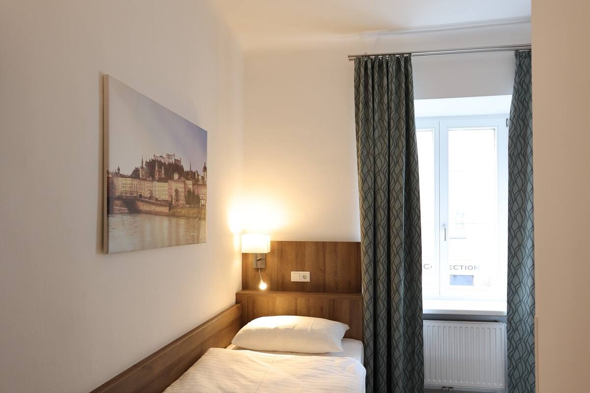 A hotel room with a bed, a nightstand, a wall-mounted lamp, a window with curtains, and a painting on the wall.