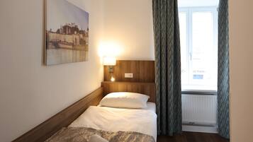 A hotel room with a bed, a nightstand, a wall-mounted lamp, a window with curtains, and a painting on the wall.