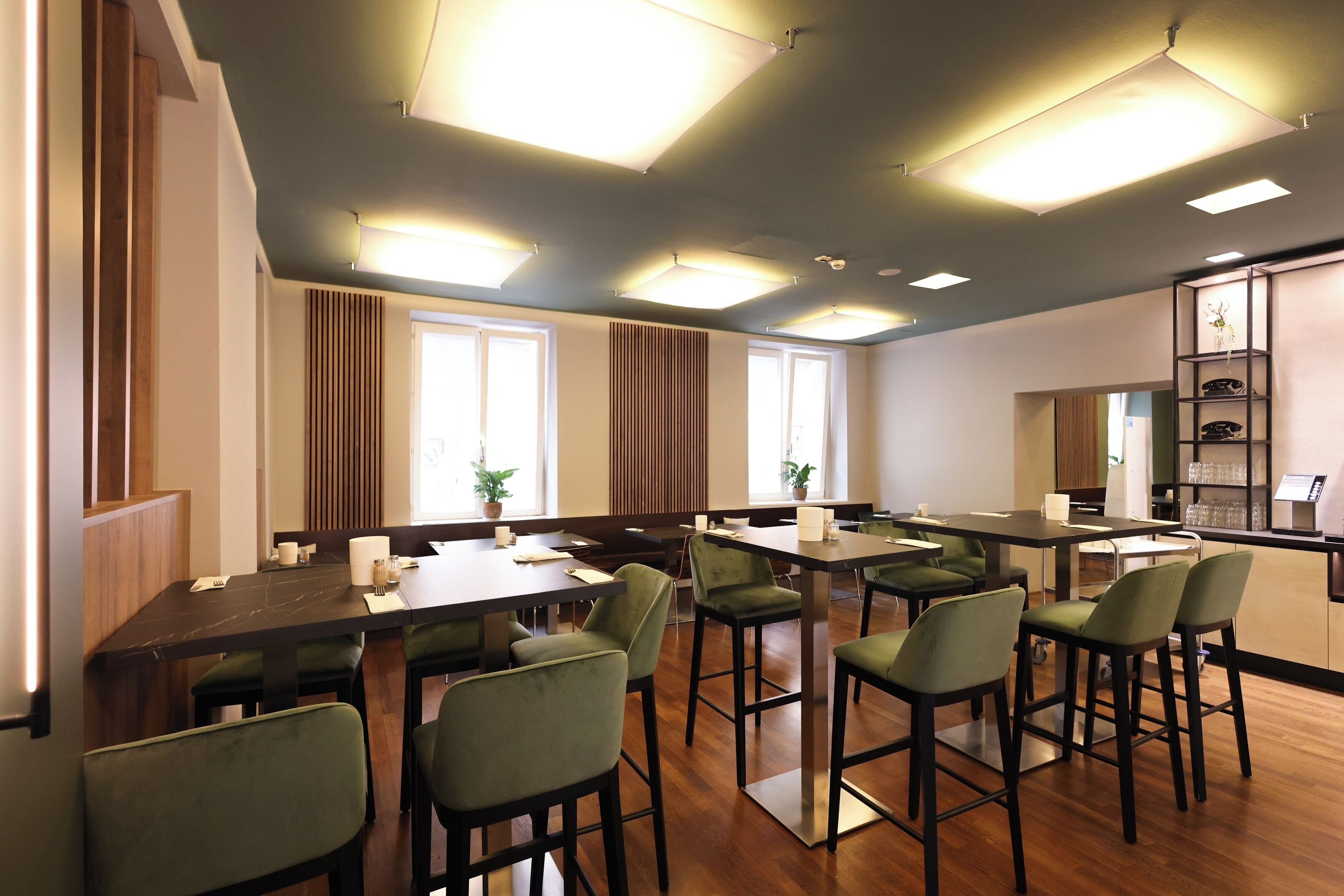 Un comedor moderno con mesas de madera y sillas verdes, ventanas amplias y una zona de bar con estanterías.