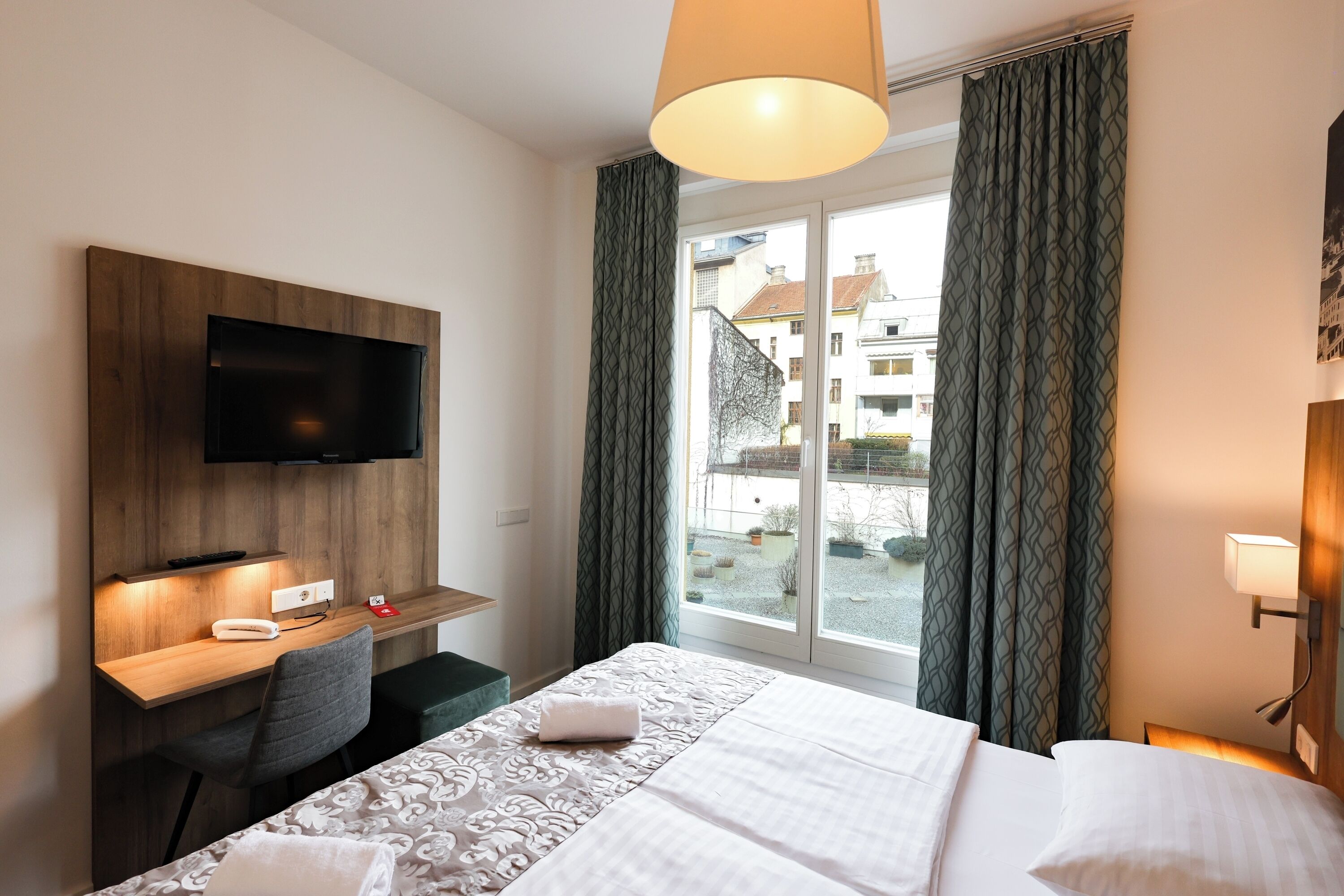 A hotel room with a bed, a desk, a TV mounted on the wall, and a window with curtains.