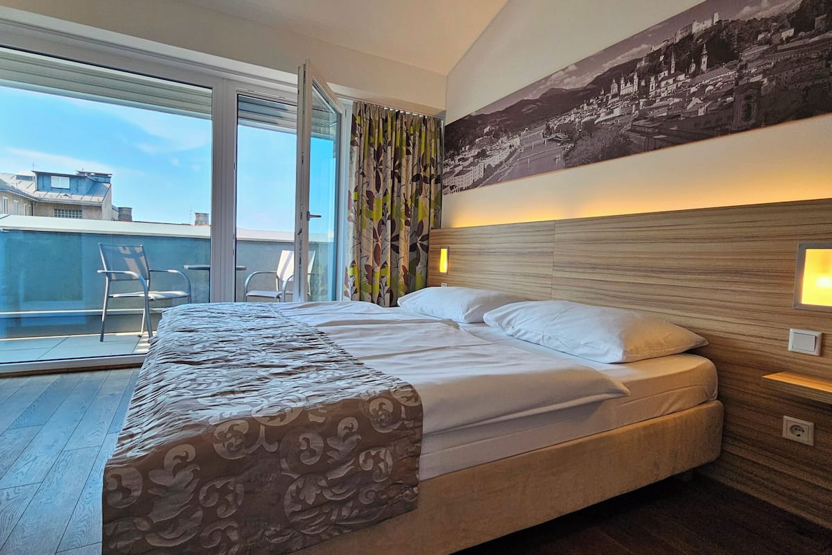 A hotel room with a large bed, a view of the city through a sliding door, and a cityscape painting on the wall.