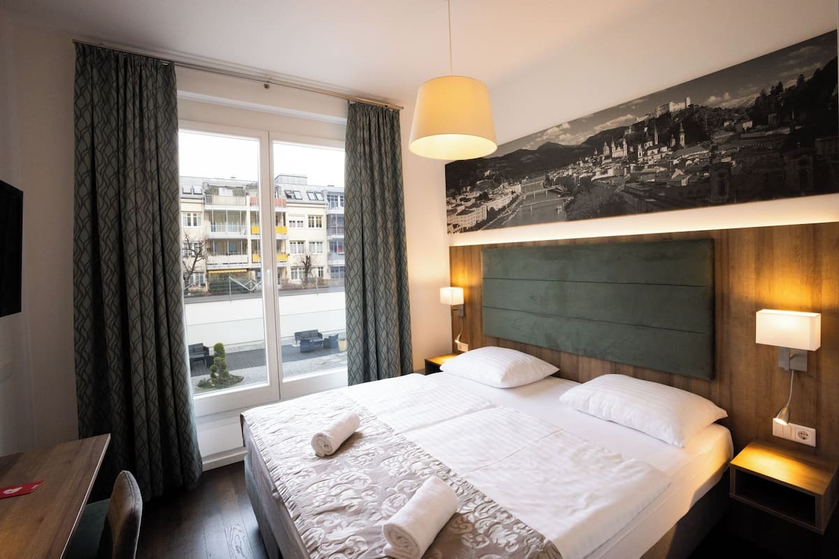 A hotel room with a large bed, a view of the city through the window, and a cityscape painting on the wall.