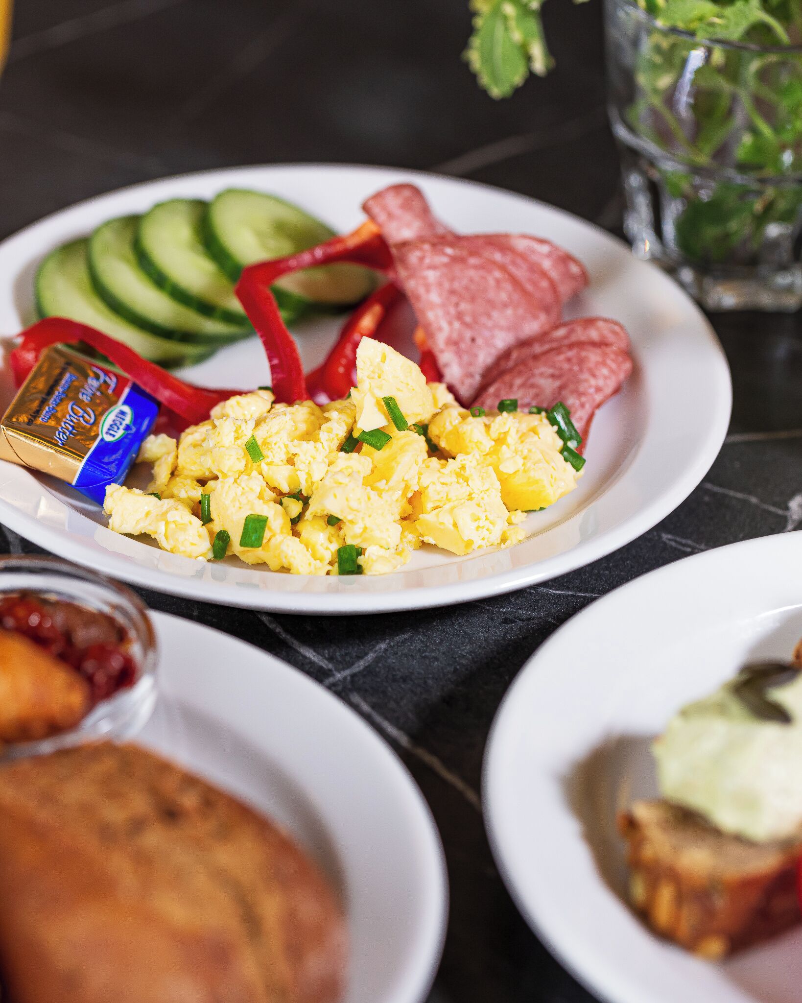 A breakfast plate with scrambled eggs, sliced cucumbers, red bell peppers, and sausages.