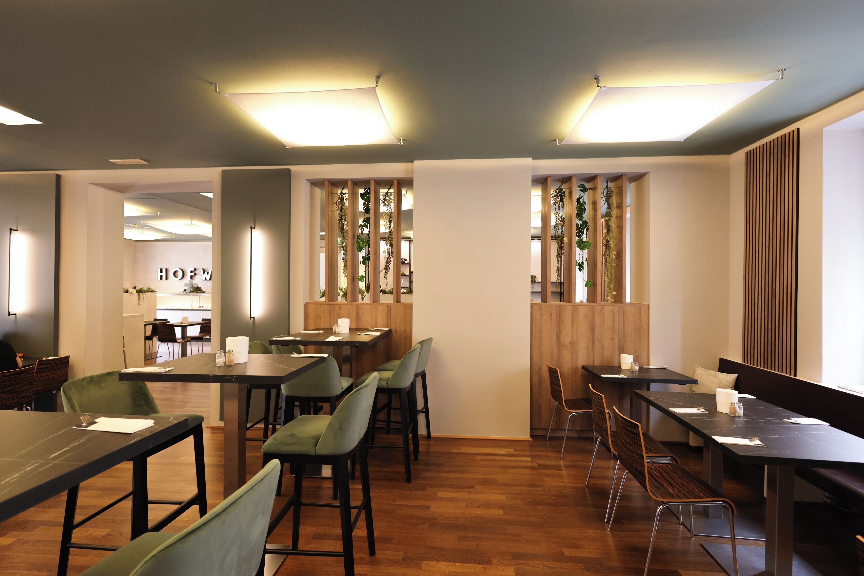 Un espace repas moderne avec des tables et des chaises en bois, une cuisine en arrière-plan et une cloison en bois agrémentée de plantes.