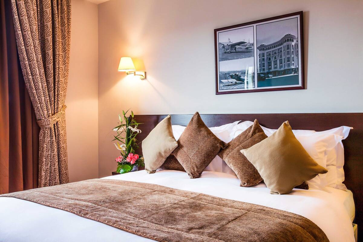 A neatly made bed with a patterned bedspread, multiple pillows, a wall-mounted lamp, and a framed picture above the bed.