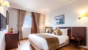 Habitación de hotel con una cama grande, una mesita de noche, un escritorio con frutero, una ventana con cortinas y lámparas de pared.