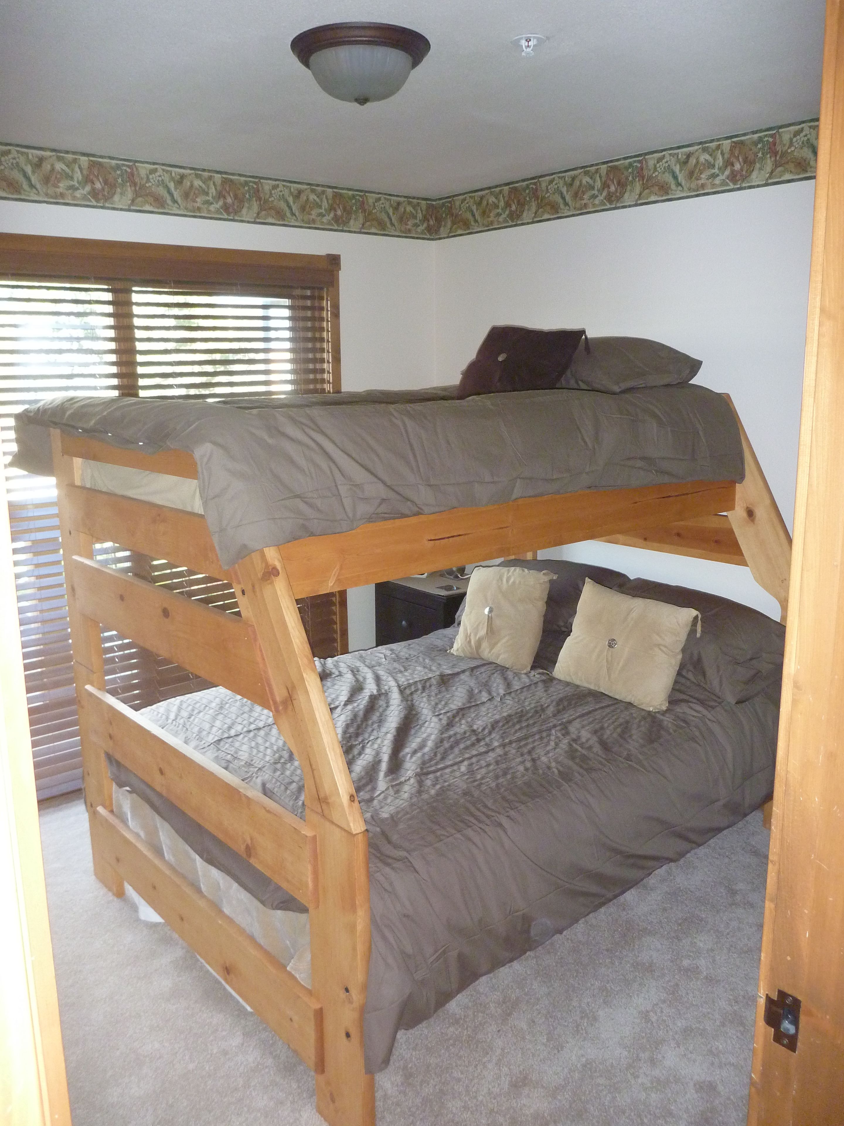 Una cama litera con estructura de madera, ropa de cama gris y una ventana con persianas.