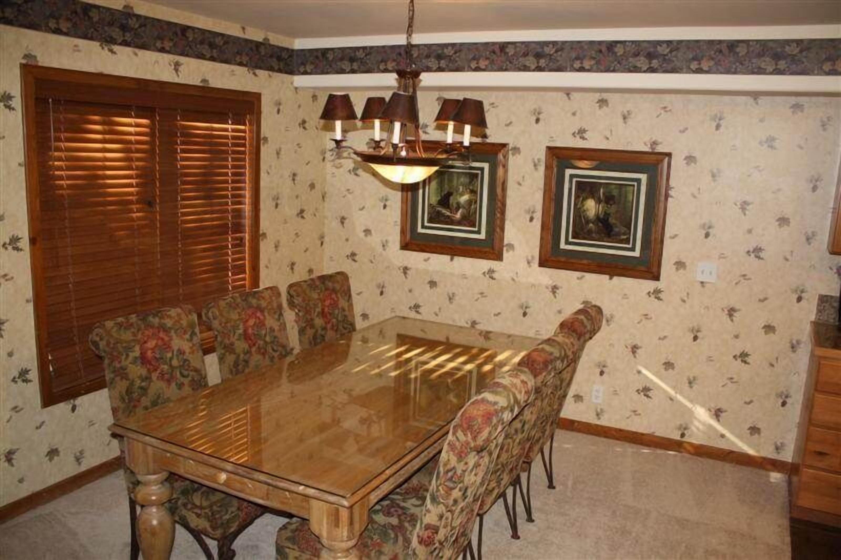 A dining room with a glass-top table, floral-patterned chairs, wooden blinds, a chandelier, and framed artwork on the wall.