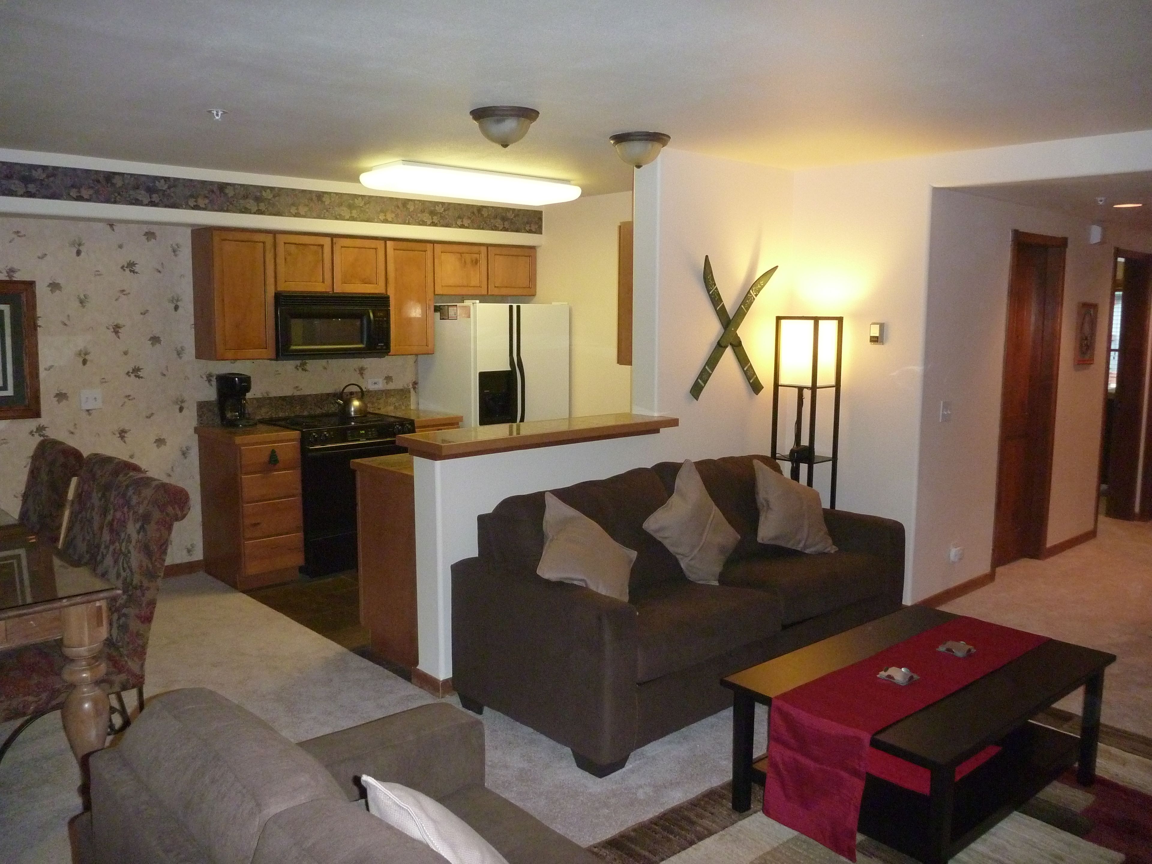 A living room with a brown sofa, a coffee table, and a kitchen area with wooden cabinets and appliances.
