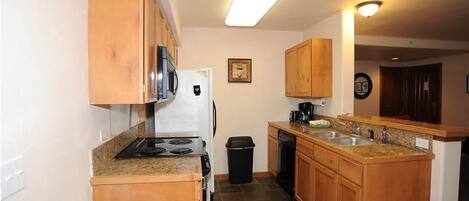 A compact kitchen with wooden cabinets, a stove, a refrigerator, a dishwasher, and a granite countertop.