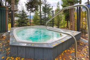 A hot tub surrounded by a wooden deck with autumn leaves, set against a backdrop of trees and a distant mountain.