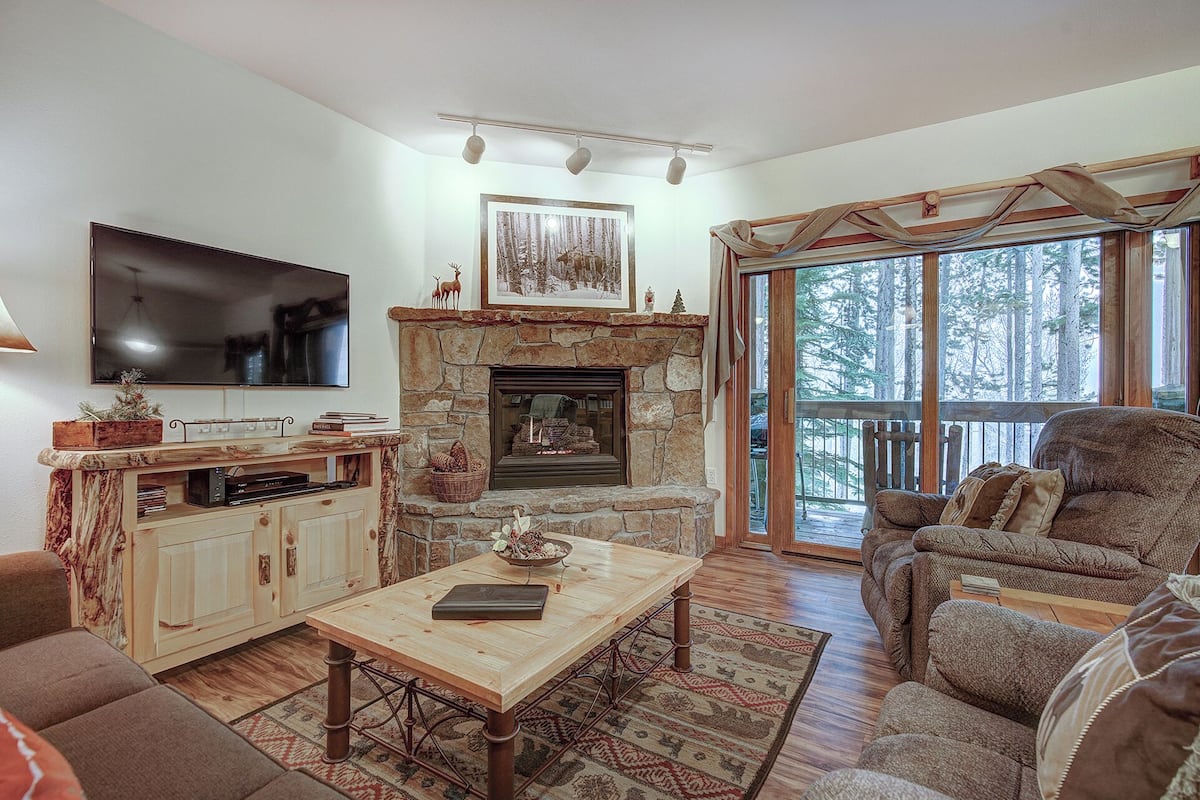 A cozy living room with a stone fireplace, a television mounted on the wall, and a large window offering a view of the forest.
