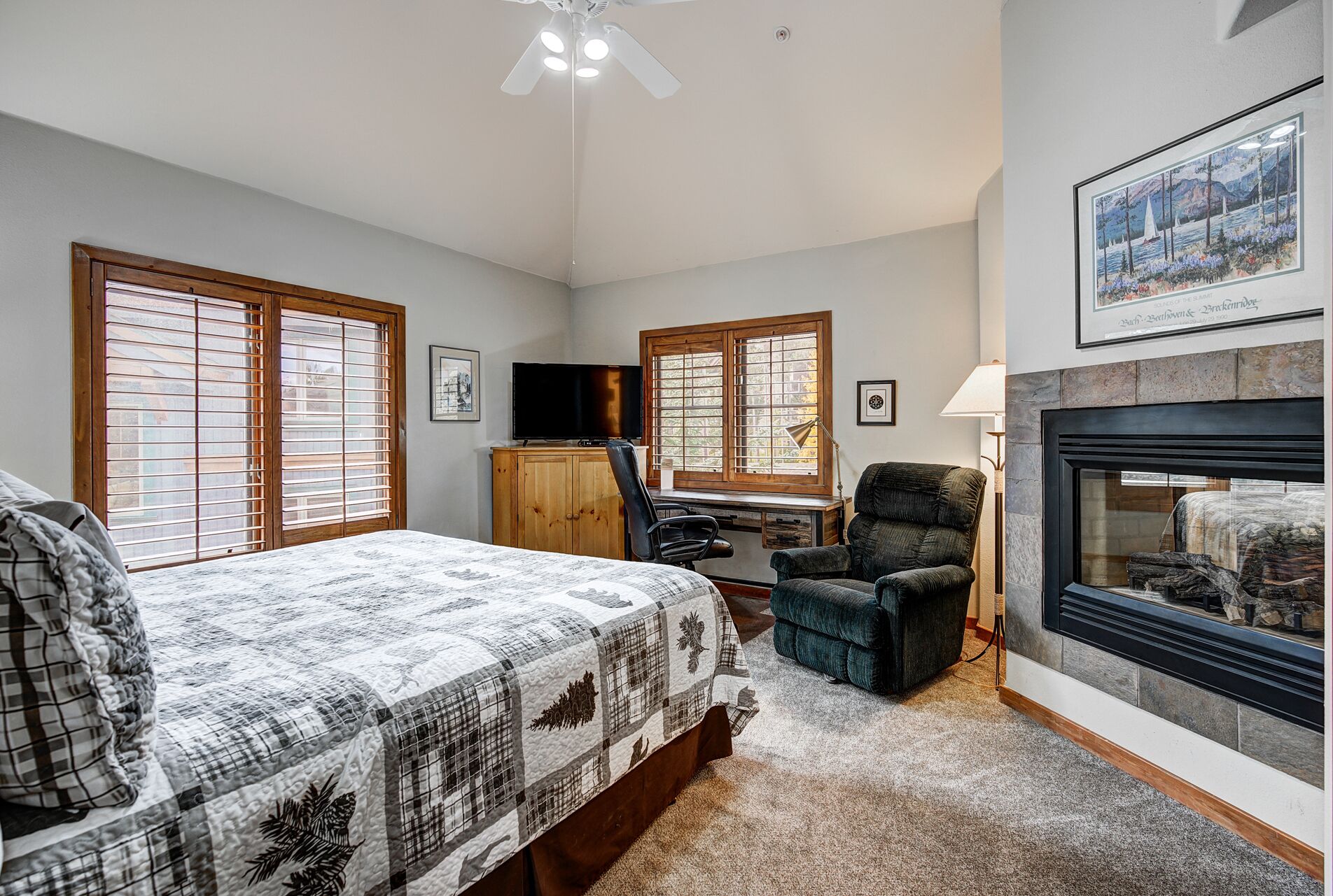 A bedroom with a bed, a desk, a chair, a fireplace, and large windows with blinds.