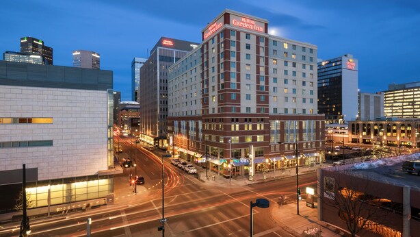 A multi-story hotel with a prominent sign for the Hilton Garden Inn, surrounded by other buildings and a busy street at dusk. - Hilton Garden Inn Denver Downtown (Denver)