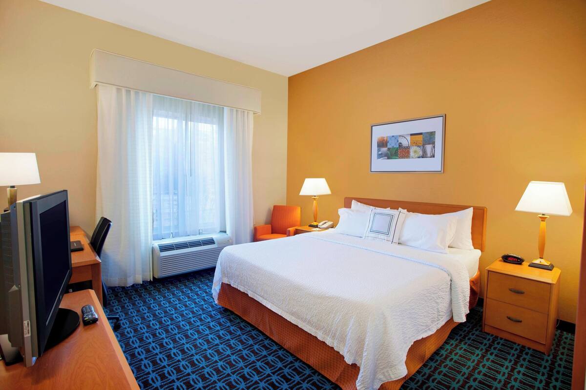 Une chambre d’hôtel avec un lit, des lampes de chevet, un bureau avec un ordinateur, une télévision et une fenêtre avec des rideaux.