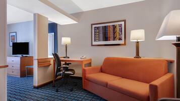 A hotel room with a blue patterned carpet, an orange sofa, a desk with a chair, a television, and two lamps.