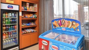Mostrador refrigerado con diversas bebidas y aperitivos, congelador con helados y postres congelados, y una ventana con cortinas.