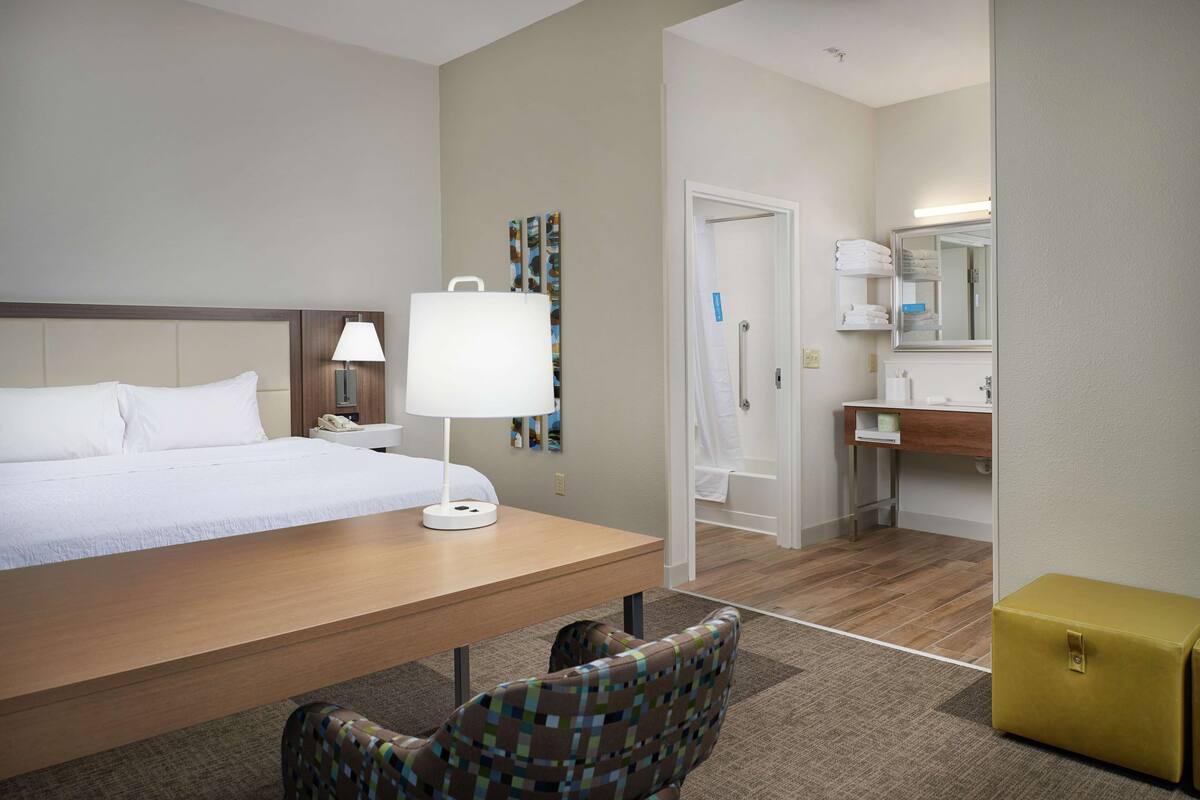 A hotel room with a bed, a desk with a lamp, a chair, a small yellow storage box, and a bathroom with a mirror and towel rack.