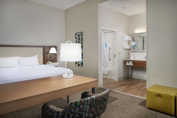 A hotel room with a bed, a desk with a lamp, a chair, a small yellow storage box, and a bathroom with a mirror and towel rack.
