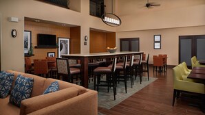A hotel lobby with a brown sofa, yellow chairs, a dining table with chairs, a TV mounted on the wall, and a ceiling fan. - Hampton Inn & Suites Rogers (Rogers)