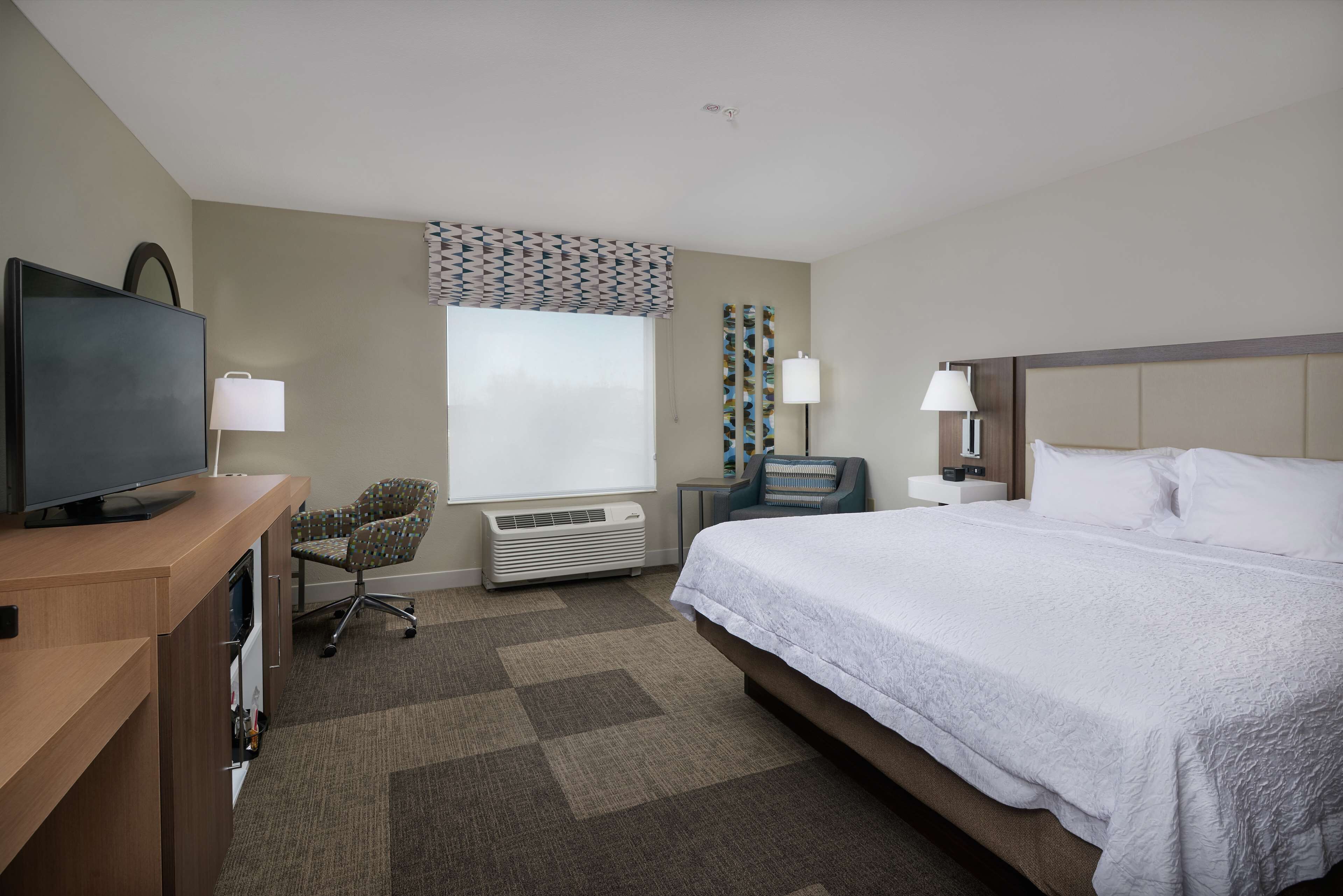 A hotel room with a bed, a desk with a television, a chair, a window with patterned curtains, and a wall-mounted lamp.