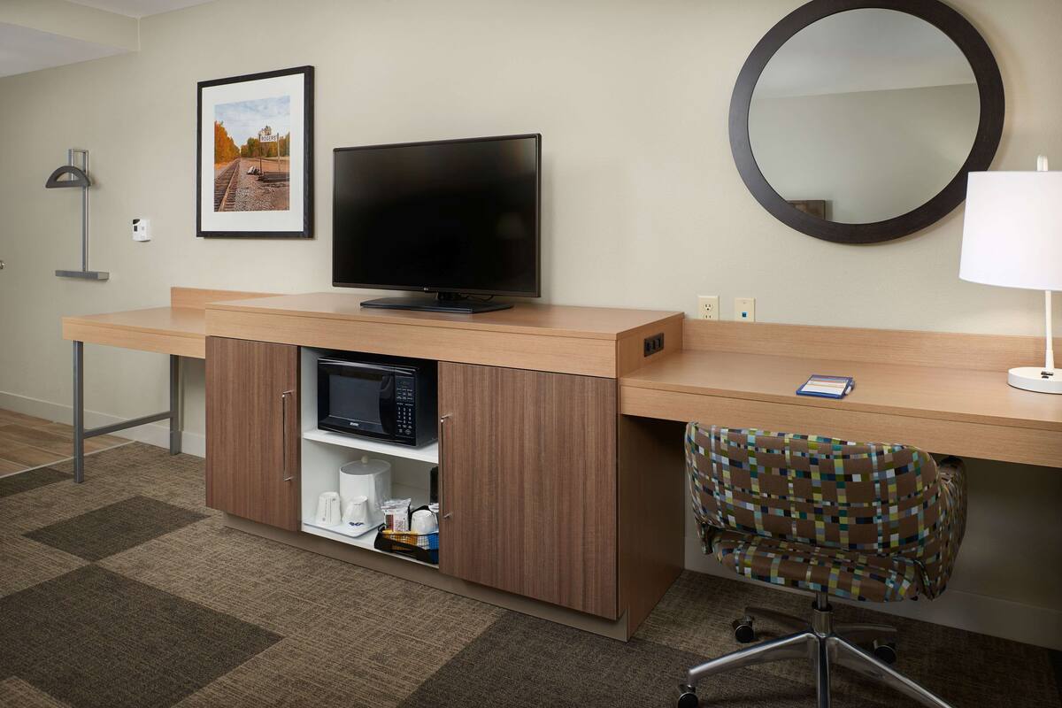 A hotel room with a wooden desk, a flat-screen TV, a microwave, a small shelf with toiletries, a patterned chair, and a round mirror.