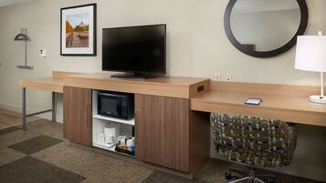 Habitación de hotel con escritorio de madera, televisor de pantalla plana, microondas, estante pequeño con artículos de aseo, silla estampada y espejo redondo.