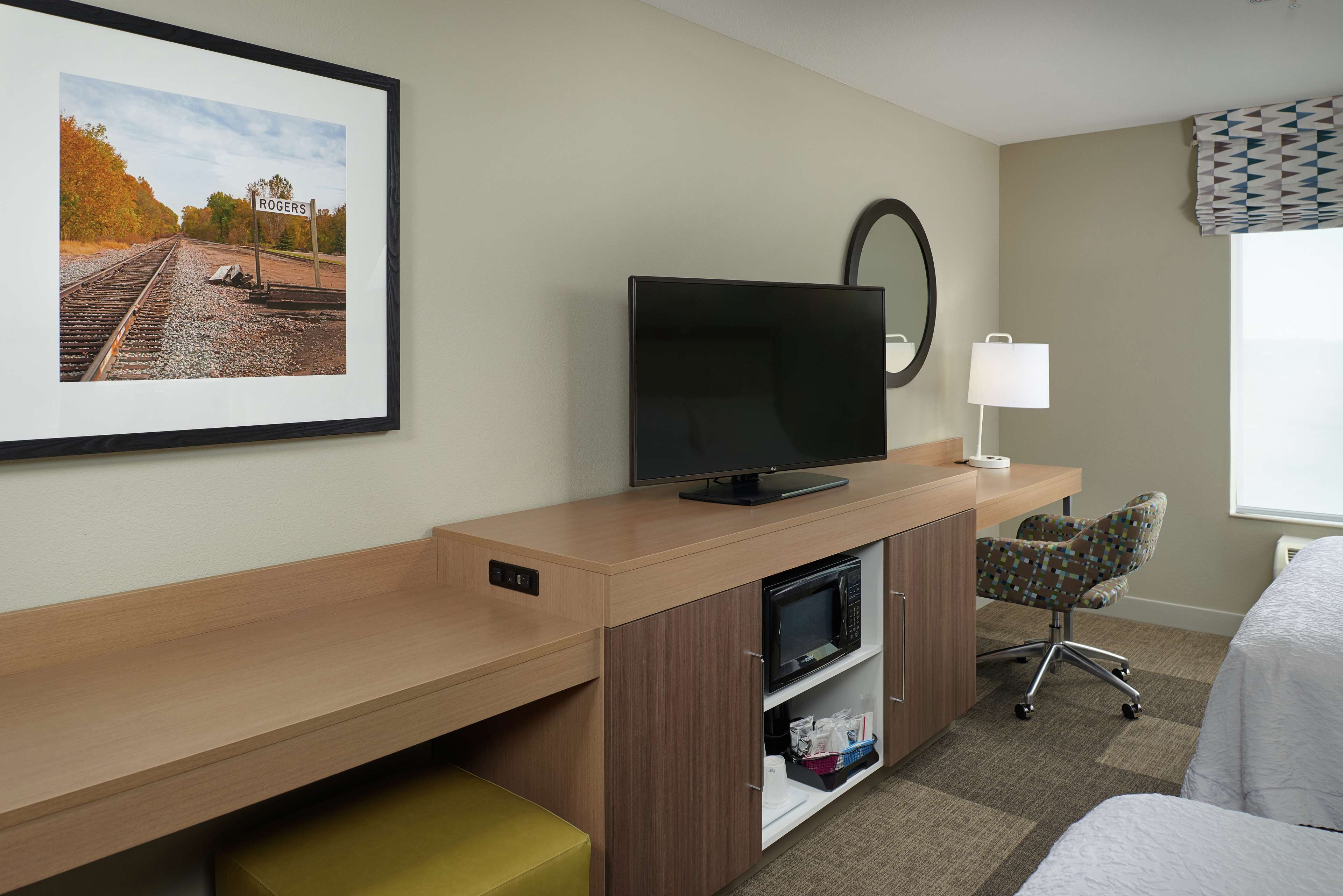 Una habitación de hotel con una cama, un escritorio con televisor, una silla, un microondas y un cuadro con una imagen de una vía de tren.