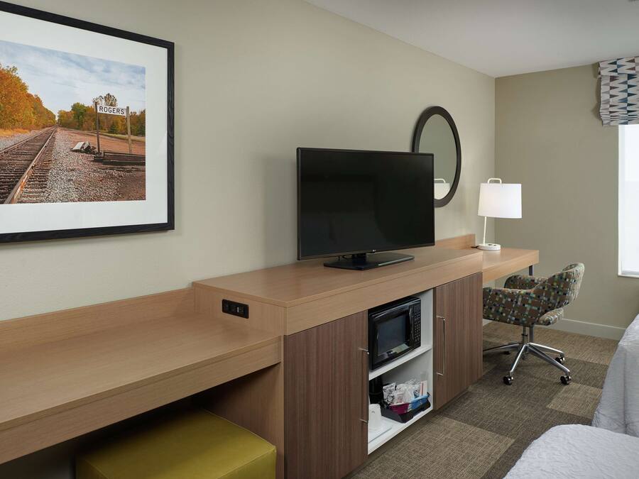 Una habitación de hotel con una cama, un escritorio con televisor, una silla, un microondas y un cuadro con una imagen de una vía de tren.