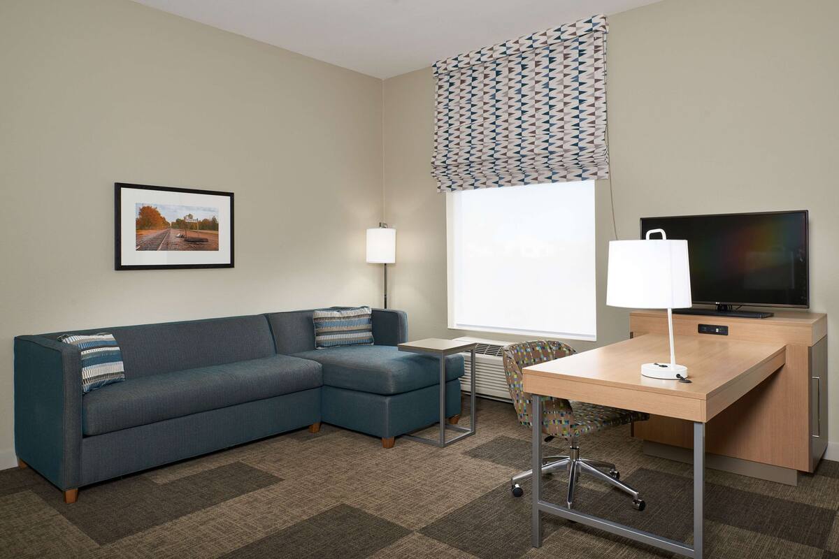 A hotel room with a sectional sofa, a desk with a lamp, a television, and a patterned window treatment.