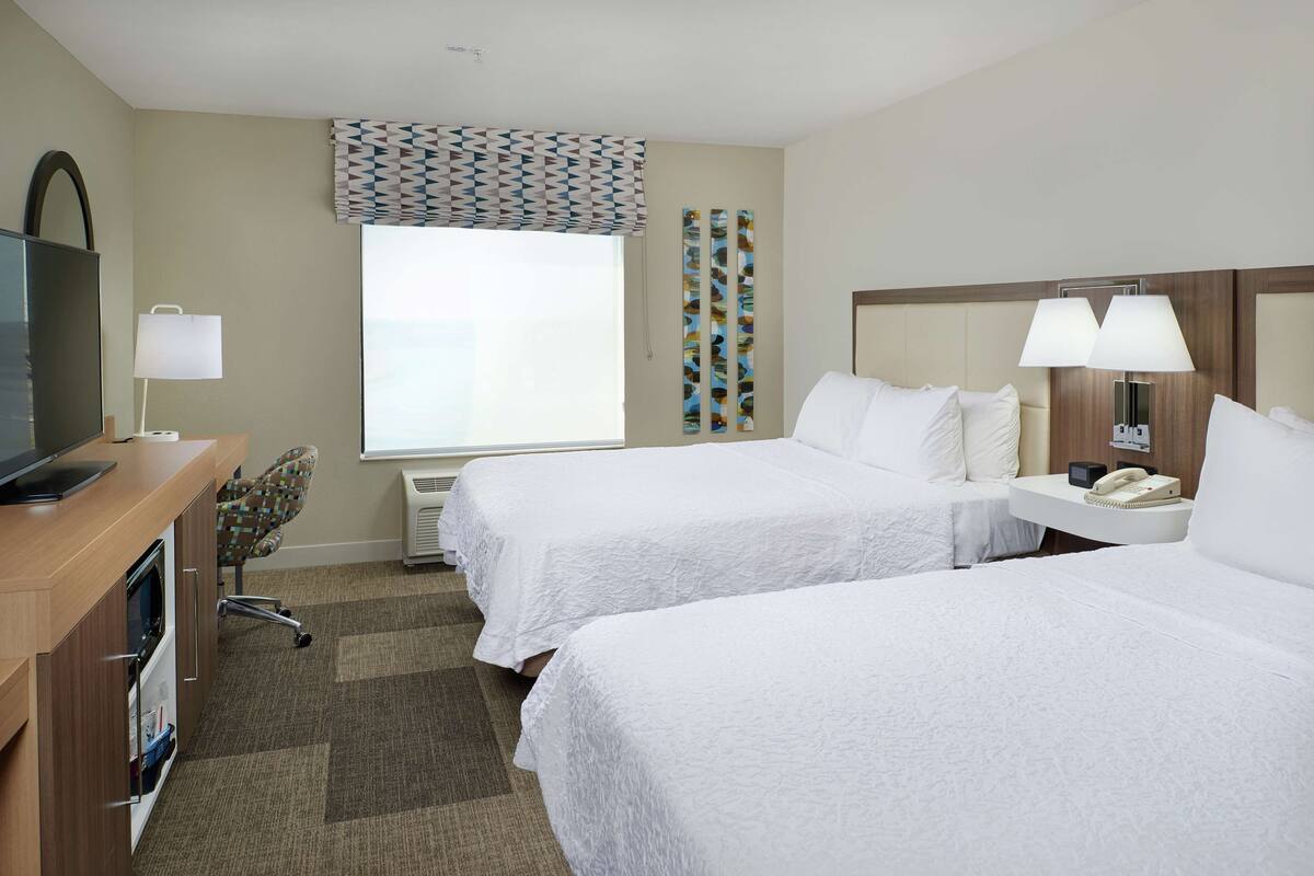 A hotel room with two beds, a desk with a computer, a chair, a TV, a window with patterned curtains, and a wall with a decorative panel.