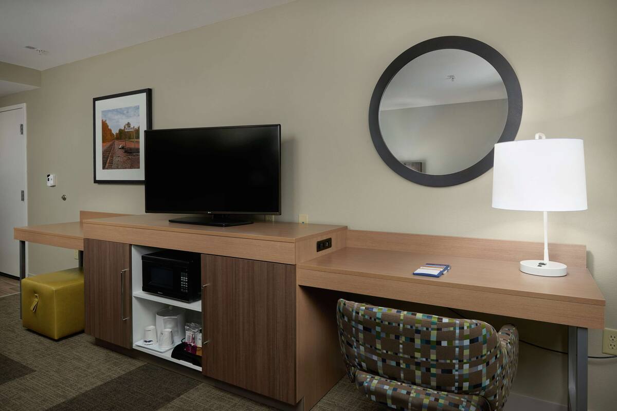 A hotel room with a wooden desk, a television, a mirror, a lamp, a patterned armchair, and a small yellow ottoman.